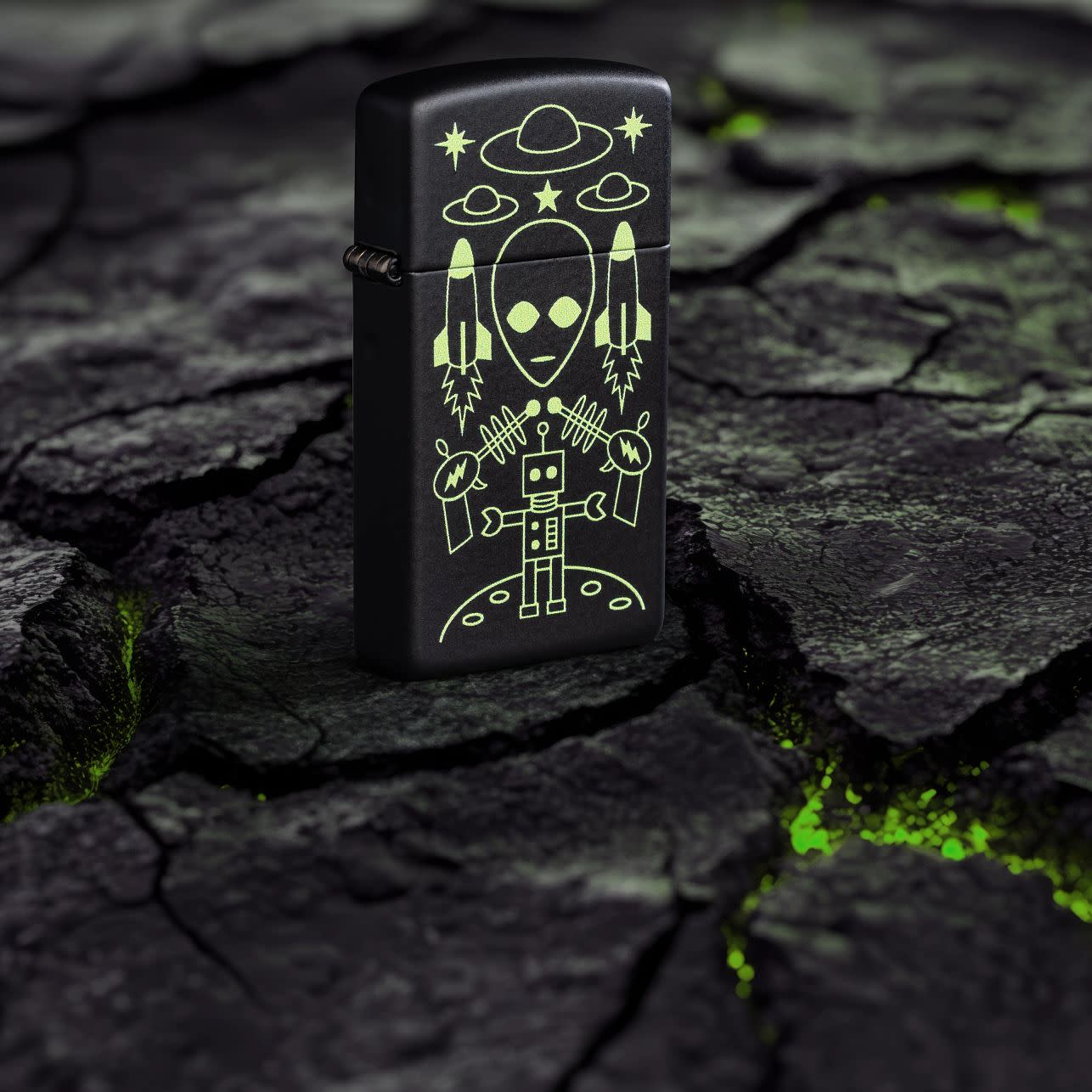 Lifestyle image of Zippo Alien Invasion Design Slim?? Black Matte Windproof Lighter standing on blacktop with green glowing cracks.