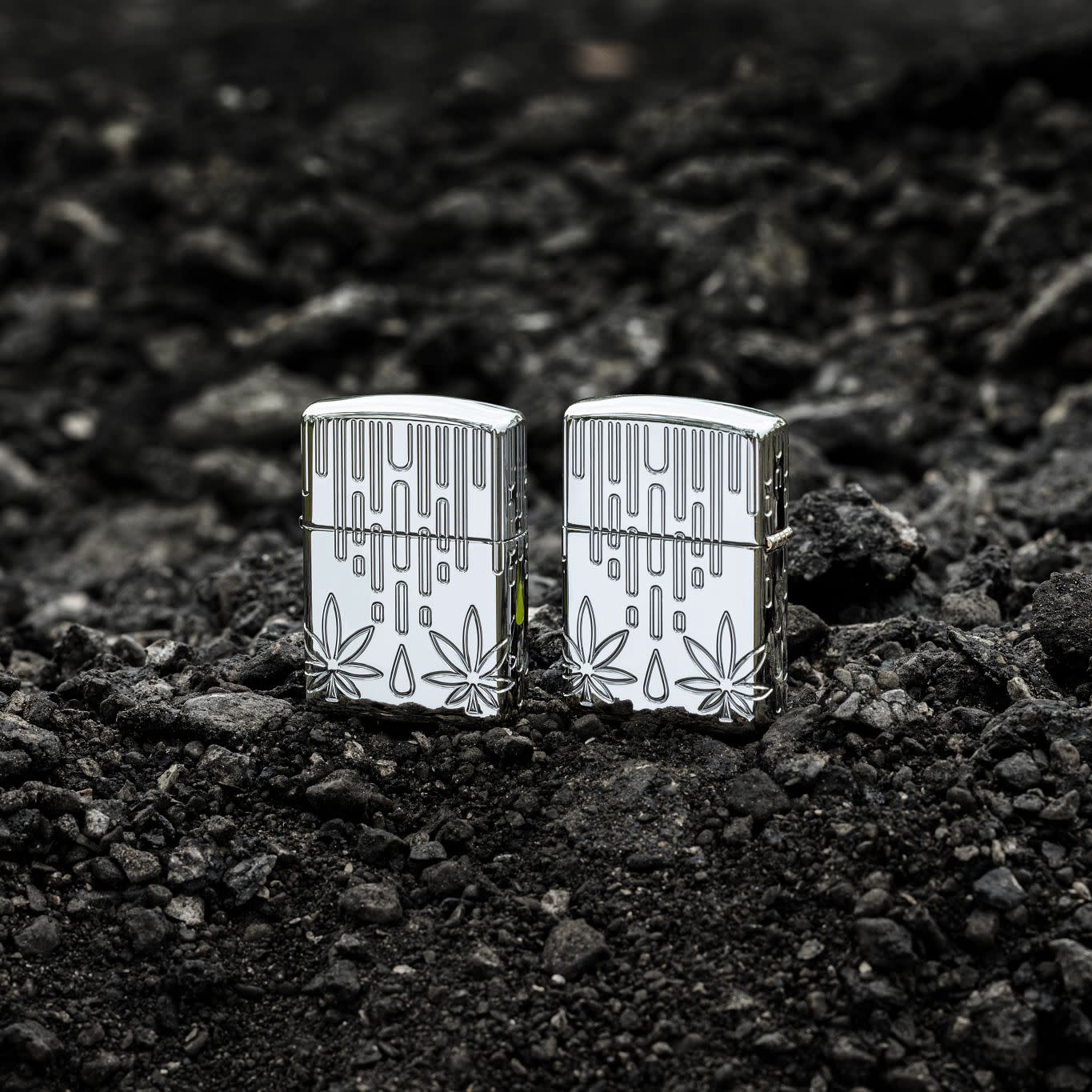 Lifestyle image of two Zippo Waxy Leaf Design Armor?? High Polish Chrome Windproof Lighters standing on dark, loose rocks; one showing the front of the lighter and the other showing the back.