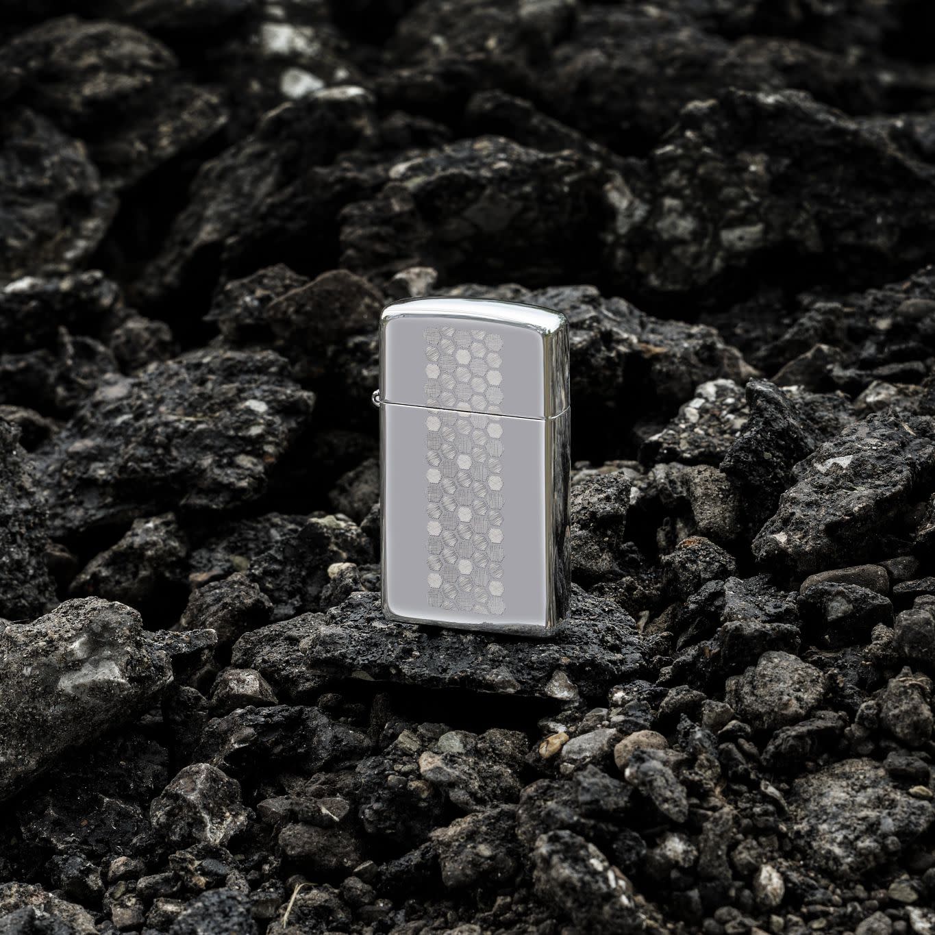Lifestyle image of Zippo Slim?? Honeycomb Design High Polish Chrome Windproof Lighter standing on dark, loose rocks.