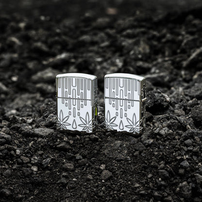 Lifestyle image of two Zippo Waxy Leaf Design Armor?? High Polish Chrome Windproof Lighters standing on dark, loose rocks; one showing the front of the lighter and the other showing the back.