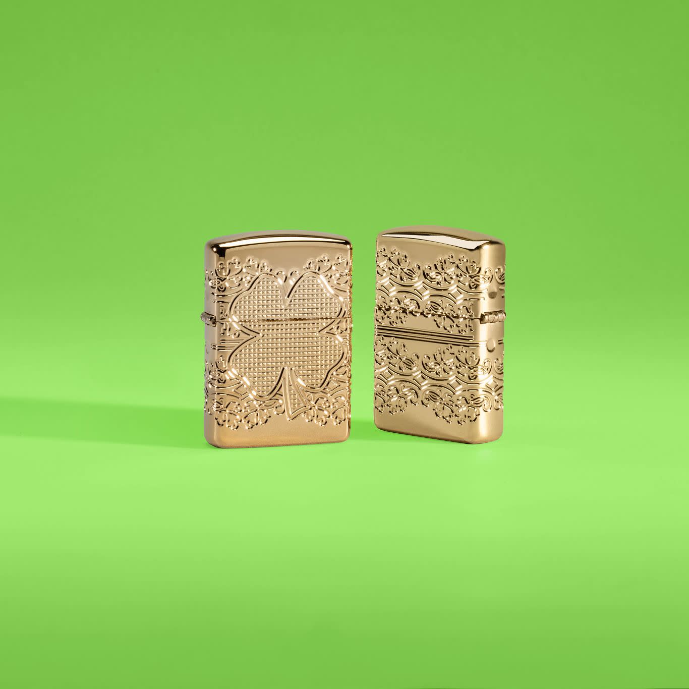 Lifestyle image of two Zippo Lucky Clover Design Armor?? High Polish Gold Plate Windproof Lighters in a lime green scene, one showing the front of the lighter and the other showing the back.